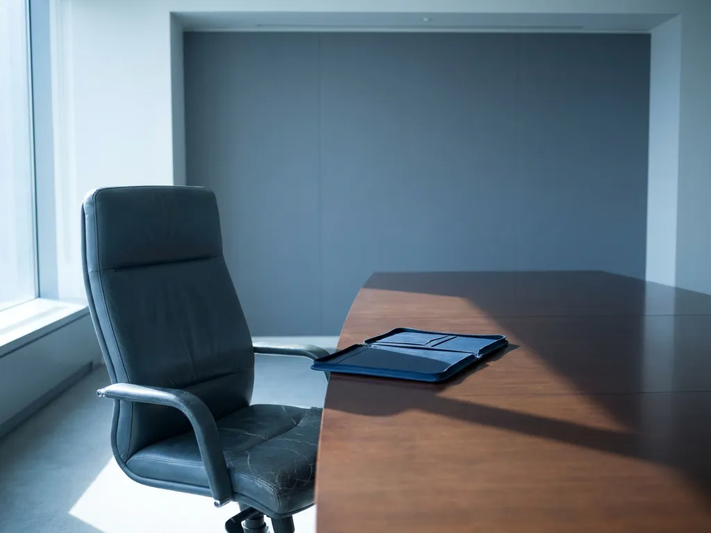 Gebarsten leren directeursstoel aan hoofd van vergadertafel met opengeslagen portfolio, lange schaduw in koel blauwgrijs licht.