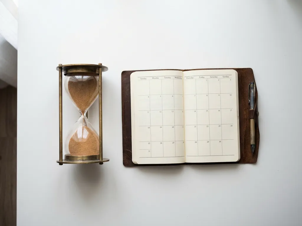 Vintage zandloper met vallend zand naast een open leren agenda op een wit bureau, minimalistisch bovenaanzicht.